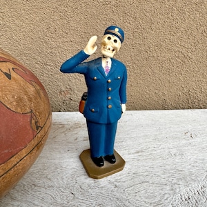 May include: A blue and white figurine of a skeleton wearing a blue uniform and a hat, saluting with its right hand. The figurine is standing on a gold base.