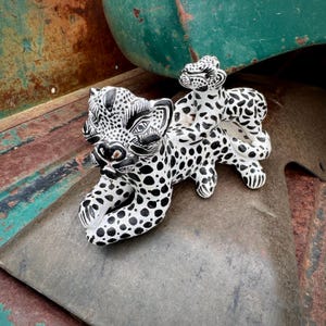 May include: A white ceramic figurine featuring a large leopard with black spots and a smaller leopard cub resting on its back. Both animals have detailed black facial markings, pointed ears, and a hand-painted appearance with a glossy finish.