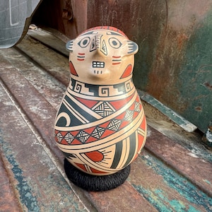 May include: A hand-painted ceramic figurine with a human-like face and geometric patterns in black, red, and white. The figurine is standing on a black base.