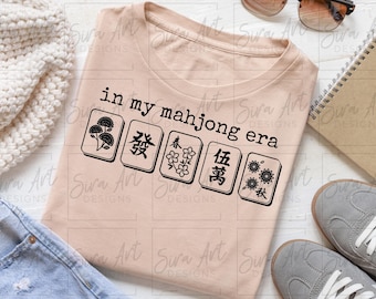 May include: A light peach-colored t-shirt with the text "in my mahjong era" and images of mahjong tiles. The shirt is surrounded by a pair of gray sneakers, sunglasses, a notebook, and a knitted cream-colored scarf.