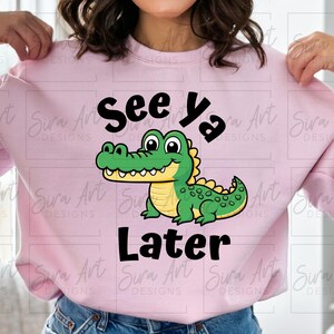 K&ouml;nnte beinhalten: Rosafarbener Pullover mit einem Comic-Alligator und dem Text "See Ya Later".