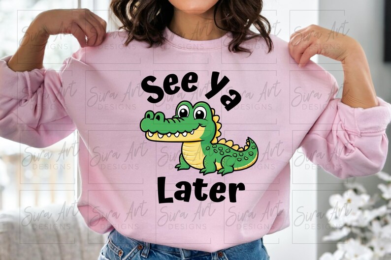 K&ouml;nnte beinhalten: Rosafarbener Pullover mit einem Comic-Alligator und dem Text "See Ya Later".