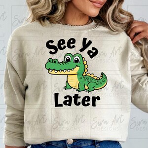 K&ouml;nnte beinhalten: Ein hellbeiger Pullover mit einem gr&uuml;nen Alligator-Cartoon-Motiv und dem Text "See ya Later" in Schwarz.