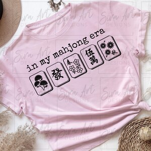 May include: A light pink t-shirt with the text "in my mahjong era" and several mahjong tiles in black. The shirt is styled with a straw hat and decorative elements.