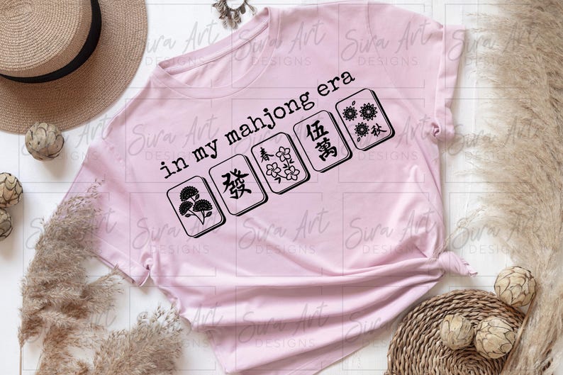 May include: A light pink t-shirt with the text "in my mahjong era" and several mahjong tiles in black. The shirt is styled with a straw hat and decorative elements.