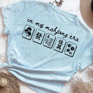 May include: Light blue t-shirt with the text "in my mahjong era" and five Mahjong tiles. The tiles feature floral and Chinese character designs. A straw hat and decorative elements are in the background.