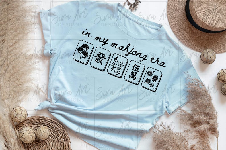 May include: Light blue t-shirt with the text "in my mahjong era" and five Mahjong tiles. The tiles feature floral and Chinese character designs. A straw hat and decorative elements are in the background.
