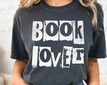 May include: Dark gray t-shirt with the words "BOOK LOVER" in a distressed white font. The shirt is a casual style, suitable for everyday wear. The design is a bold statement for book enthusiasts.
