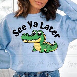 K&ouml;nnte beinhalten: Hellblauer Pullover mit einem Comic-Alligator und dem Text "See ya Later" in Schwarz.