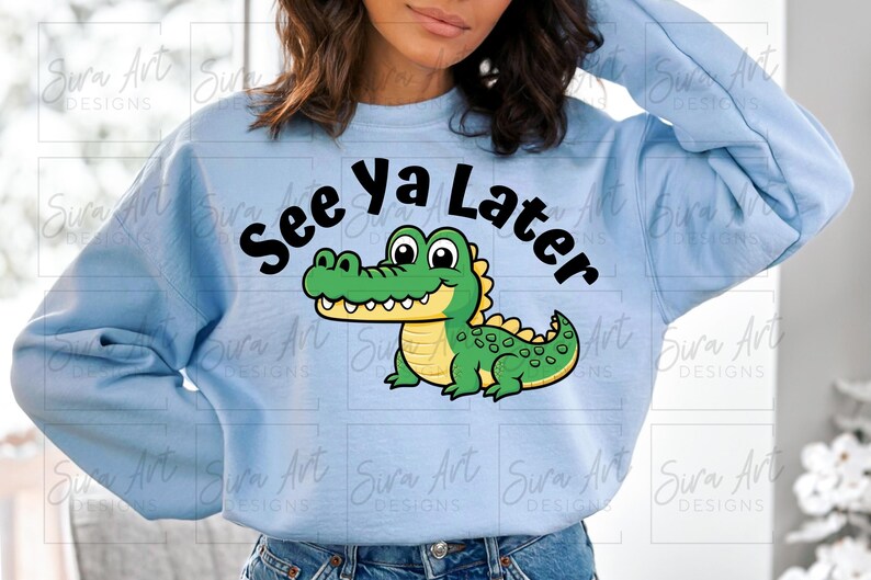 K&ouml;nnte beinhalten: Hellblauer Pullover mit einem Comic-Alligator und dem Text "See ya Later" in Schwarz.