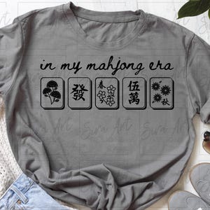 May include: Gray t-shirt with the text "in my mahjong era" above five black and white mahjong tiles. The tiles feature various symbols. The shirt is displayed on a white surface with accessories.