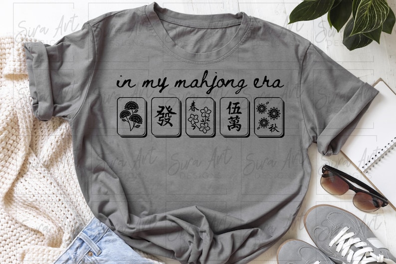 May include: Gray t-shirt with the text "in my mahjong era" above five black and white mahjong tiles. The tiles feature various symbols. The shirt is displayed on a white surface with accessories.