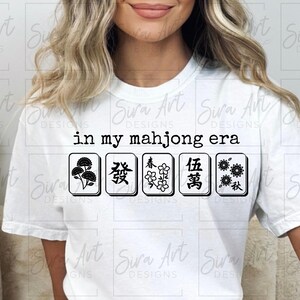 May include: White t-shirt with the phrase "in my mahjong era" and five black and white mahjong tiles. The tiles feature various symbols. A casual, comfortable top for game enthusiasts.
