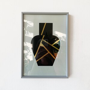 May include: A framed print of a black vase with gold geometric lines on a light blue background.