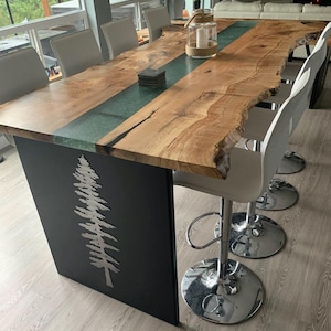 May include: A live edge wood dining table with a green epoxy resin inlay. The table has a black metal base with a tree design. The table is surrounded by white bar stools with chrome bases.