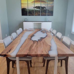 Peut inclure: Une grande table à manger en bois avec un incrustation de résine époxy blanche. La table a un piétement en métal blanc et est entourée de chaises blanches.