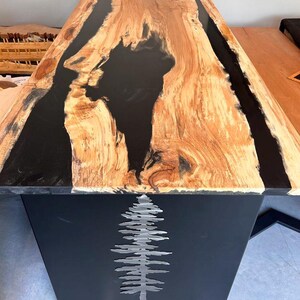 May include: A wooden table top with a black epoxy resin river running through it. The table top is light brown with a natural wood grain. The table has a black metal base with a silver metal tree design.
