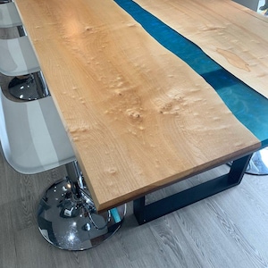 May include: A large wooden table with a blue epoxy river down the centre. The table has a light brown wood grain finish and a black metal base. The table is approximately 183 cm long and 91 cm wide.