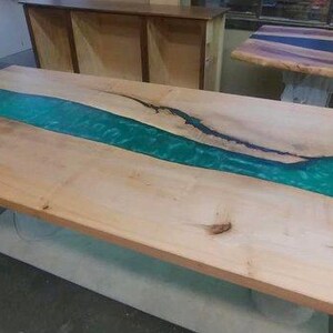 May include: A large, rectangular wooden table with a teal epoxy resin river feature. The table is made of light-coloured wood with a natural grain. The table is supported by a white base. The table's length is approximately 200 cm.