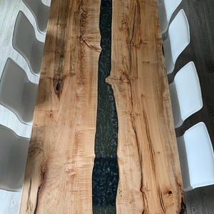 May include: A long, wooden dining table with a natural wood finish and a black epoxy river running down the centre. The table is surrounded by white chairs.