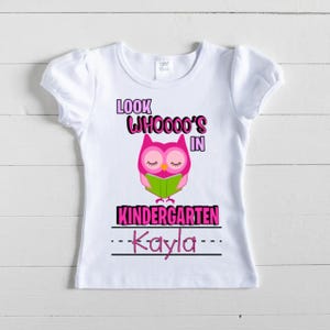 May include: White short-sleeved t-shirt with a pink and green owl graphic. The shirt has the text "LOOK WHOOOO'S IN KINDERGARTEN" and the name "Kayla" in pink. The shirt is designed for children.