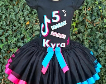 May include: Black tulle tutu skirt with pink and blue ribbon trim. A black t-shirt with a pink and blue TikTok logo, the number 5, and the text "It's My Birthday Kyra" is worn over the skirt.