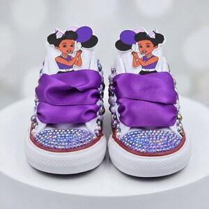 Op de afbeelding: Een paar witte hoge sneakers met paarse satijnen lintveters, neuzen bezet met iriserende kristallen en zijkanten versierd met paarse en witte parels. Op de tong is een uitgesneden cartoonfiguur bevestigd die paarse ballonnen vasthoudt.
