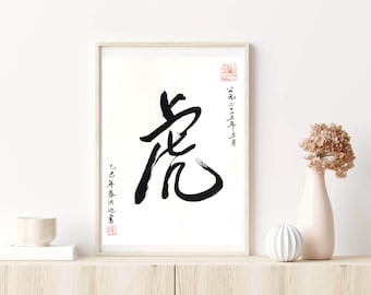 May include: A framed artwork with a striking black calligraphy character on a white backdrop. The character is the main element, accompanied by vertical text and red stamps. The artwork is displayed on a light wood surface, with a vase and decorative sphere.