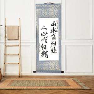 May include: A decorative wall scroll featuring Japanese calligraphy in black ink on a white panel. The scroll has a blue and gold patterned border and a wooden handle. A wooden ladder with a towel and a rug are in the background.