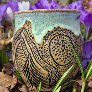 Puede incluir: Una taza de cerámica con un esmalte verde claro y un diseño marrón y negro de montañas y árboles. La taza está sentada sobre una cama de hierba verde y hojas marrones.