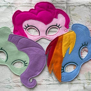 May include: Three felt masks of My Little Pony characters. The masks are pink, purple and blue with rainbow accents. Each mask has a hole for the eyes and a felt design of a pony's face.