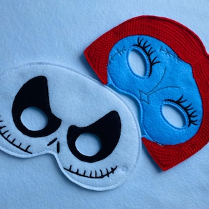 May include: Two felt Halloween masks, one white with black details and the other blue with red hair and black eyelashes.