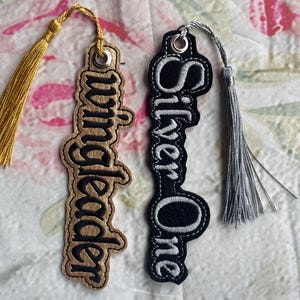 May include: Two fabric bookmarks with tassels. One bookmark is gold with black embroidery that says "Wunderleder". The other bookmark is black with white embroidery that says "Silver One".