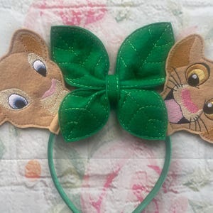 May include: A headband with two tan lion cub faces and a green leaf-shaped bow. The lion cubs have embroidered details and the headband is light green. The cubs have blue and yellow eyes.