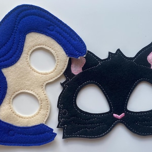 May include: Two felt masks, one blue with a white face and pink flower, the other black with pink ears and a white face.