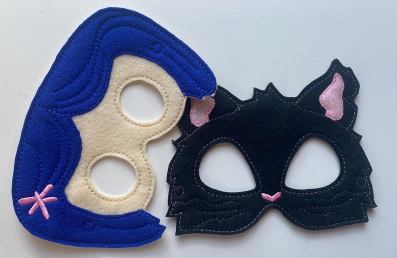 May include: Two felt masks, one blue with a white face and pink flower, the other black with pink ears and a white face.