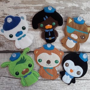 May include: A collection of felt animal character patches, including a polar bear, a dark-skinned character, a cat with an eye patch, a green alien, a monkey, and a penguin. Each character wears a blue hat and collar.