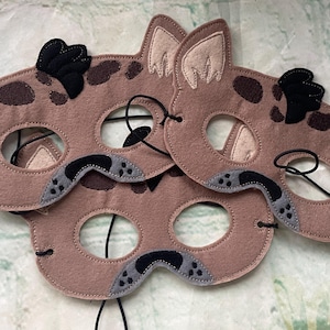 May include: Three tan felt animal masks featuring dark brown spots, pointed ears with cream-colored inner sections, black tufts of hair, and gray snout details with black stitching. Each mask includes black elastic cords for securing around the head.