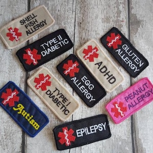 May include: A collection of embroidered medical alert patches in various colors. Each patch features a red medical symbol and text indicating conditions such as "Shellfish Allergy," "Type II Diabetic," and "Peanut Allergy."