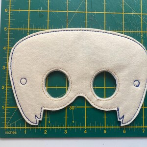 May include: A white felt mask with two eye holes and a stitched design around the edges. The mask has a blue stitching around the edges and two small circles on the sides.