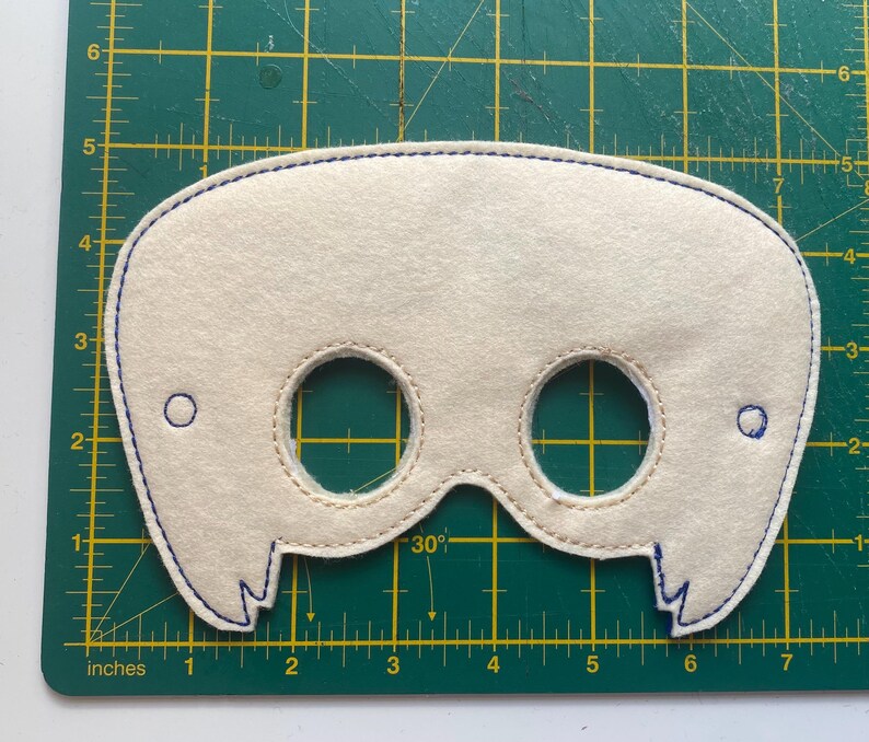 May include: A white felt mask with two eye holes and a stitched design around the edges. The mask has a blue stitching around the edges and two small circles on the sides.