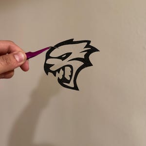 May include: A black and white decal of a snarling animal head with a pointed ear and a wide open mouth. The decal is being held up by tweezers.