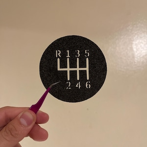 Puede incluir: Un círculo negro con números y líneas blancas que representan el patrón de cambio de marchas de un coche. Los números son R, 1, 3, 5, 2, 4 y 6.