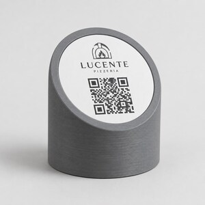 Pode incluir: Um expositor de mesa cilíndrico cinzento com uma inserção circular branca. A inserção apresenta o texto "LUCENTE PIZZERIA" acima de um código QR e um gráfico de forno de pizza. O expositor tem um topo inclinado e uma superfície texturizada e canelada.