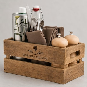 Puede incluir: Una caja de madera rectangular con el texto OSTERIA MILANO y un icono de copa de vino grabados en el frente. La caja contiene un recipiente de aceite de metal alto, una botella de vidrio, cubiertos de plata, servilletas de tela marrón y dos molinillos.