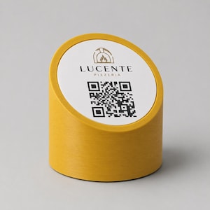 Può includere: Un espositore da tavolo cilindrico giallo con una parte superiore circolare bianca. La parte superiore presenta il testo "LUCENTE PIZZERIA" e un codice QR. L'espositore ha una parte superiore inclinata e una superficie strutturata.