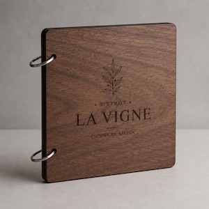 Puede incluir: Una funda de menú cuadrada de madera marrón oscuro con dos anillas de metal plateado en el lado izquierdo. El frente presenta un gráfico botánico grabado con láser sobre el texto BISTROT LA VIGNE y CUISINE DE SAISON debajo.