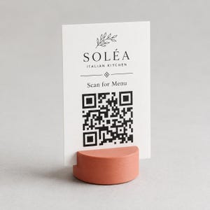 May include: A white rectangular card with black text reading SOLÉA ITALIAN KITCHEN and Scan for Menu, featuring a line drawing of a leafy branch and a QR code. The card is held upright by a semi-circular, terracotta-colored base with a grooved texture.
