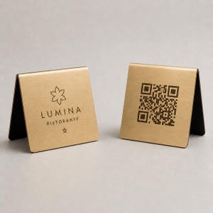 Op de afbeelding: Twee goudkleurige metalen tafelschilden. Eén bord toont de tekst "LUMINA RISTORANTE" met een bloemmotief en een ster. Het andere bord toont een QR-code. Beide borden hebben een zwart interieur.