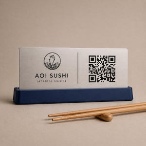 Op de afbeelding: Een geborsteld metalen bord voor "AOI SUSHI JAPANSE KEUKEN" met een QR-code en een logo van een vis boven golven. Het bord staat in een donkerblauwe houder. Twee houten eetstokjes rusten op een houten eetstokjeshouder.